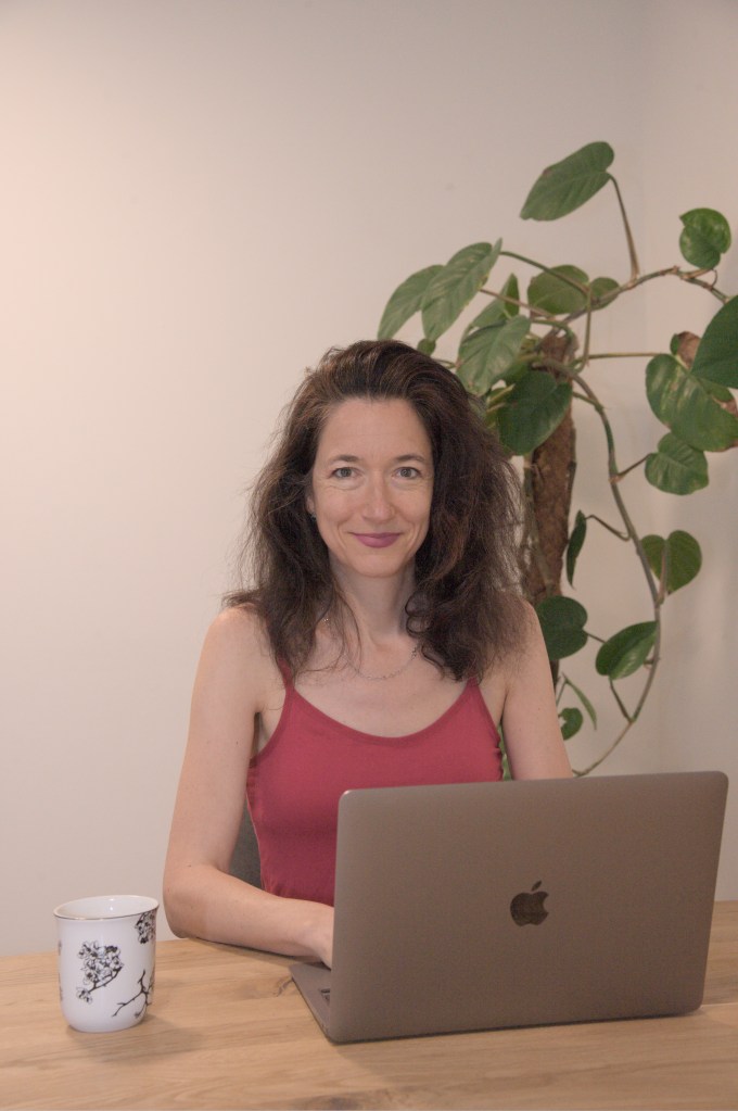 A woman sitting at a wooden table, smiling while working on a laptop, with a plant in the background and a white mug beside her.